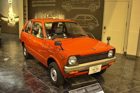 Orange small three-door hatchback with round headlamps and wing mirrors on its front wings, on a shiny museum floor before a wall of line drawings. Opens a larger view