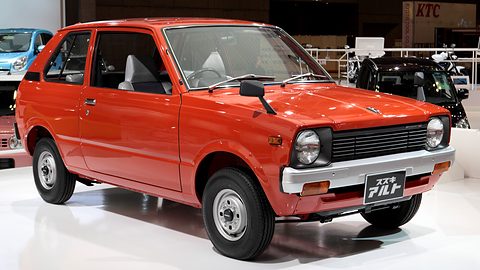 Red-orange small three-door hatchback with round headlamps, a black grille and a white bumper, on a white show floor. Opens a larger view