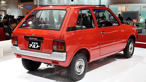 Rear quarter of a red-orange small three-door hatchback with a white rear bumper, on a white stand with visitors behind. Opens a larger view