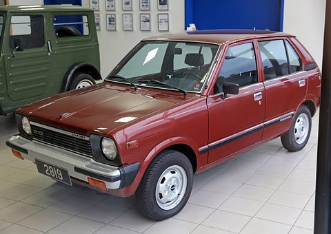 Dark red small five-door hatchback with round headlamps and a black plate, on a tiled showroom floor, a green Jimny behind. Opens a larger view
