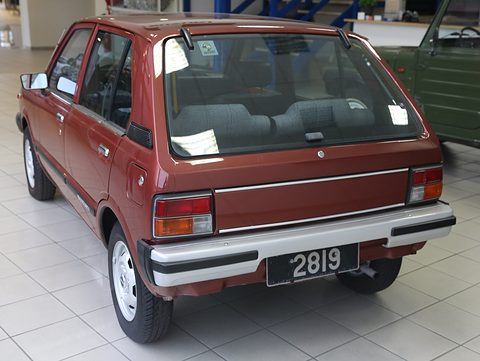 Rear quarter of a dark red small five-door hatchback with a chrome-trimmed bumper, on a white tiled showroom floor. Opens a larger view