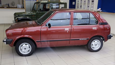 Side view of a dark red small five-door hatchback with a Suzuki side stripe, a green 4x4 behind it in a showroom. Opens a larger view