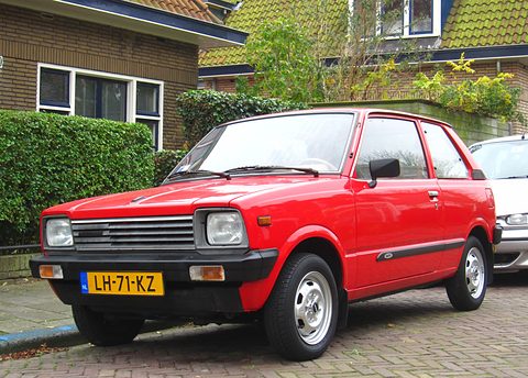 Red small three-door hatchback with square headlamps, a slatted black grille and a Dutch plate, parked on a brick street by a hedge. Opens a larger view