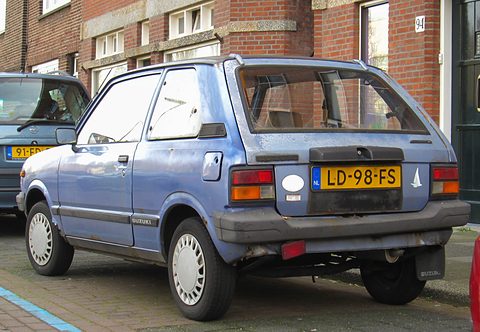 Rear quarter of a faded pale blue small three-door hatchback with a yellow Dutch plate, parked by a brick house. Opens a larger view