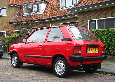 Rear quarter of a red small three-door hatchback with a black rear bumper and tail lamps, parked on a brick street by houses. Opens a larger view
