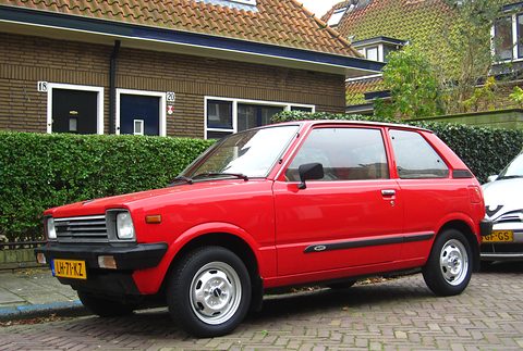 Side view of a red small three-door hatchback with a black rubbing strip and silver wheels, in front of brick houses and a hedge. Opens a larger view