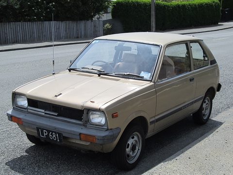 Beige small three-door hatchback with square headlamps and a black New Zealand plate, parked at a kerb by a hedge. Opens a larger view