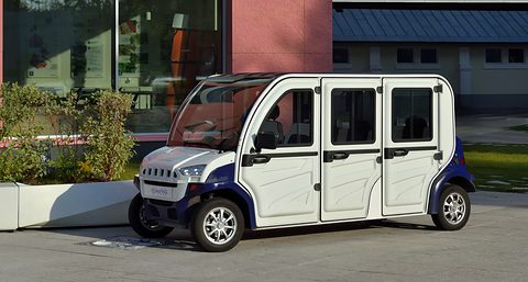 White and dark blue six-door electric shuttle with large windows parked on tarmac by shrubs and a building. Opens a larger view