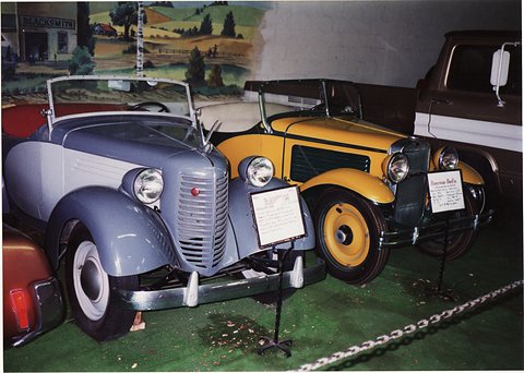 A pale blue-grey open car and a yellow and black roadster side by side on green carpet in a display. Opens a larger view