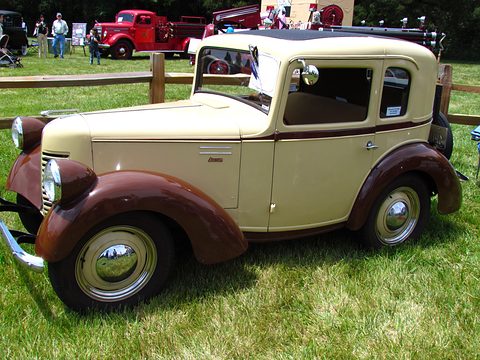 A small cream coupé with brown wings and white-wall tyres parked on grass by a wooden fence. Opens a larger view
