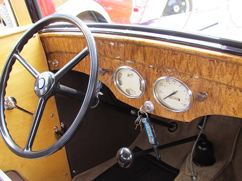 A steering wheel in front of a wood-grain painted dashboard with two round dials and a key. Opens a larger view