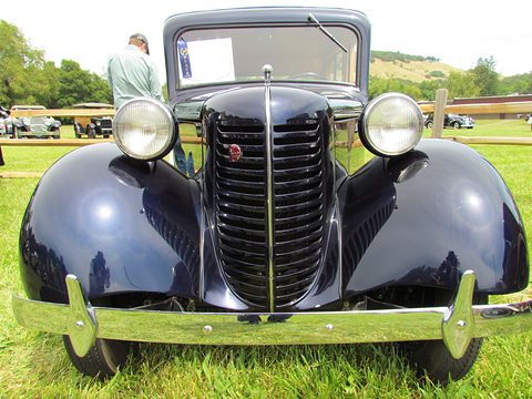 Head-on view of a black car with a tall ribbed grille, round headlamps on the wings and a chrome bumper. Opens a larger view