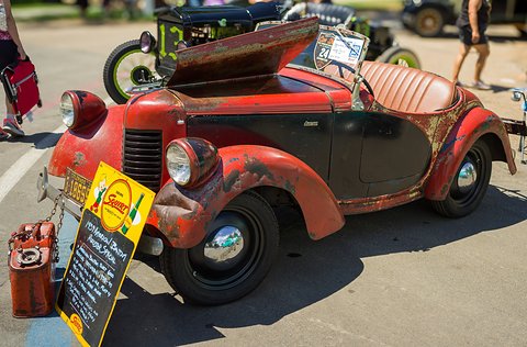 A weathered red and cream roadster with its bonnet up and a show board in front of it, on tarmac. Opens a larger view