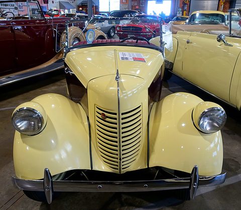 Head-on view of a pale yellow roadster with a tall ribbed grille and headlamps faired into its wings. Opens a larger view