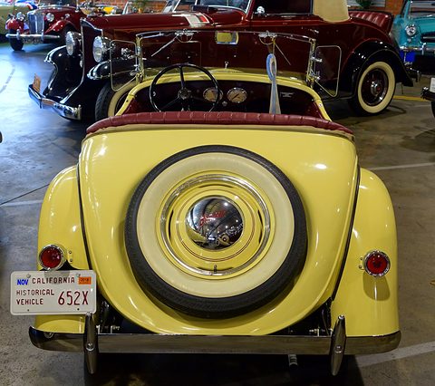 The rear of a yellow roadster with a white-walled spare wheel and a California historic vehicle plate. Opens a larger view