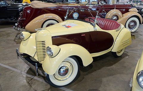 A cream roadster with a maroon side panel and red seats among other cars in a museum hall. Opens a larger view
