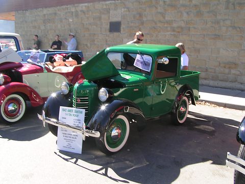 A small green pick-up with its bonnet open and display cards on the windscreen, parked before a stone wall. Opens a larger view