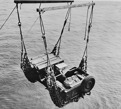 A small military car hanging in a rope net from a crane arm above dark water. Opens a larger view