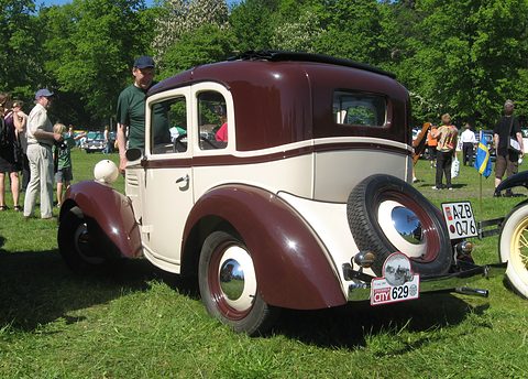 The rear of a small two-tone coupé with a spare wheel on its back, parked on grass with people beyond. Opens a larger view