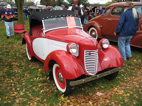 A red roadster with white side panels, white-wall tyres and a black hood on leaf-strewn grass, people around. Opens a larger view