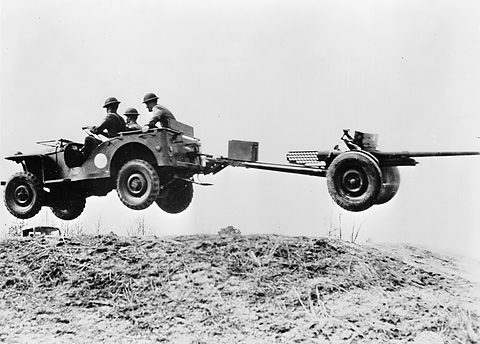 A small open military car with three soldiers aboard airborne over a mound, towing a light gun.