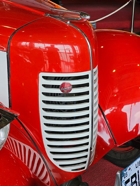 Close view of a red curved bonnet with a tall chrome grille of vertical bars and a small badge. Opens a larger view