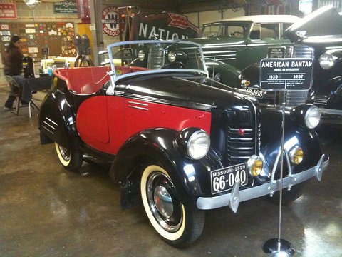 A black open car with a red side panel, white-wall tyres and a display sign on a stand, in a hall. Opens a larger view