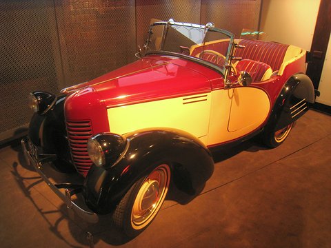 A red open car with a yellow side panel and red leather seats under warm museum light. Opens a larger view