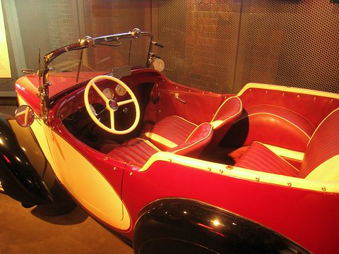 Looking over the side of a red and yellow open car at red leather seats and a cream steering wheel. Opens a larger view
