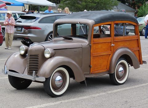 A small station wagon with a grey bonnet and wings, a wooden body and white-wall tyres in a car park. Opens a larger view