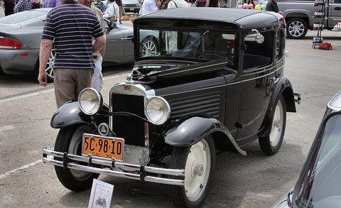 A small black two-seat coupé with cream wheels, large headlamps and bonnet louvres in a car park, a man beside it. Opens a larger view