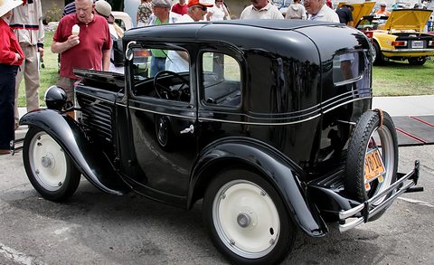 The rear quarter of a small black coupé with cream disc wheels and a rear-mounted spare, visitors behind. Opens a larger view