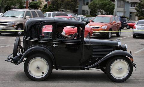 Side view of a small black coupé with cream wheels, dwarfed by the modern cars parked behind it.