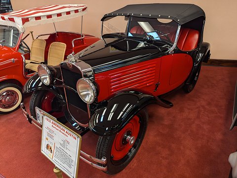 A small red open car with black wings and a black hood up, a display card before it, on red carpet. Opens a larger view