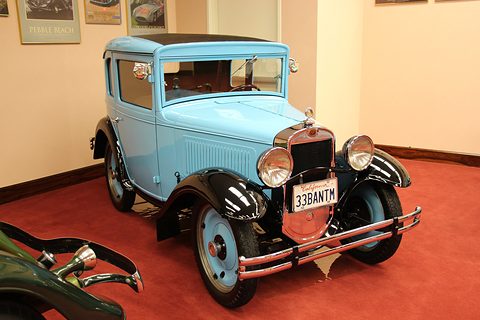 A small pale blue coupé with black wings, white-wall tyres and a chrome bumper on red carpet in a museum. Opens a larger view