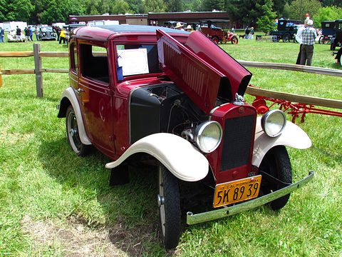 A small maroon coupé with white wings, its bonnet side raised to show the engine, parked on grass by a fence. Opens a larger view