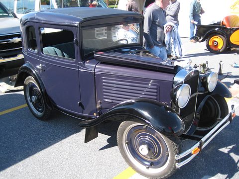 A small purple coupé with black wings and white-wall tyres in a show field, visitors behind. Opens a larger view