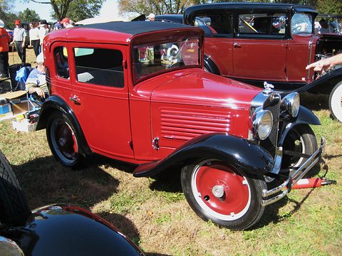 A small red coupé with black wings and red wheels on grass, other cars and visitors behind. Opens a larger view
