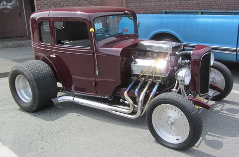 A maroon small coupé body on a low chassis with fat rear tyres, chrome wheels and an open engine at the front. Opens a larger view