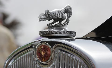 A chrome rooster mascot on a radiator cap above a round red enamel badge and a chrome grille shell. Opens a larger view