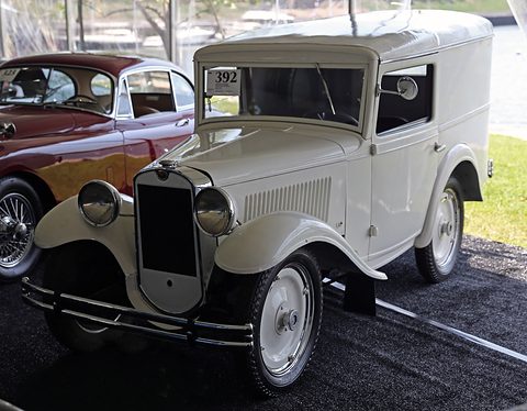 A small white panel van with a narrow grille, black wings and white-wall tyres under a tent. Opens a larger view
