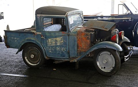 A small blue pick-up with faded, rusting paint and old lettering on its door, under a white tent. Opens a larger view