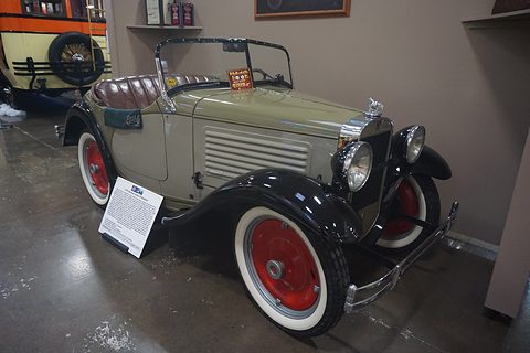 A small grey-green roadster with black wings, red wheels and a display card in a museum hall. Opens a larger view