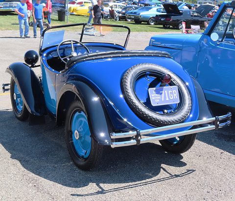 The rounded blue tail of a small roadster with a spare wheel on the back and a chrome rear bumper. Opens a larger view