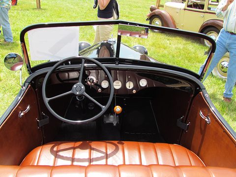 Looking into an open car over tan pleated leather seats to a black steering wheel and a row of small dials. Opens a larger view