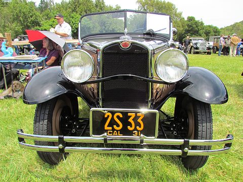 Head-on view of a small black roadster with a tall radiator, two large headlamps and a yellow California plate, on grass. Opens a larger view