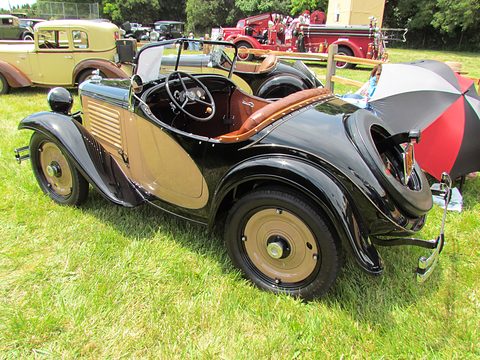 A small black roadster with tan side panels, cream wheels and a rumble seat open, parked on grass under umbrellas. Opens a larger view