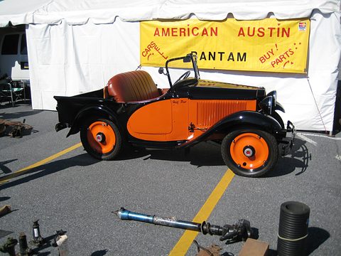 Side view of a small orange open car with orange wheels in front of a white tent with a yellow club banner. Opens a larger view