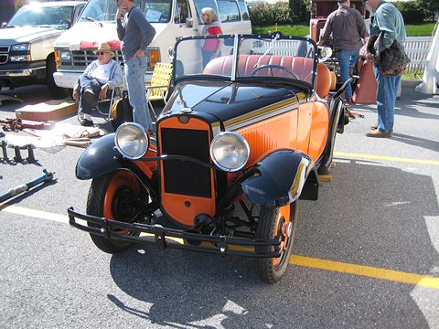 A small orange roadster with black wings and its hood folded in a busy show field, people and parts around it. Opens a larger view