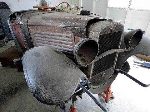 The rusty front of a small car with peeling paint, its wings lifted away from the chassis, in a workshop. Opens a larger view